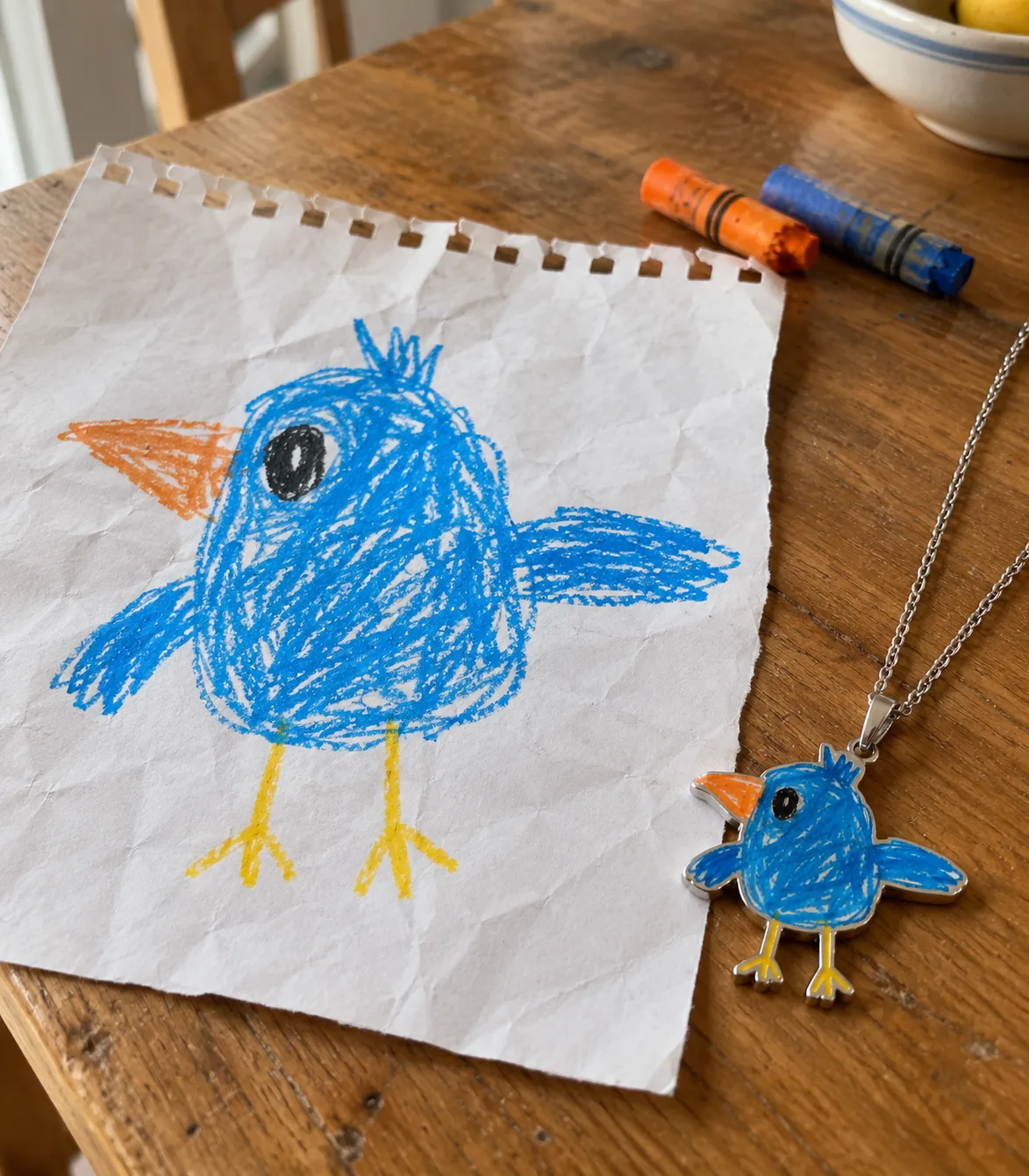 A child's blue crayon bird drawing on notebook paper beside the matching bluebird pendant necklace