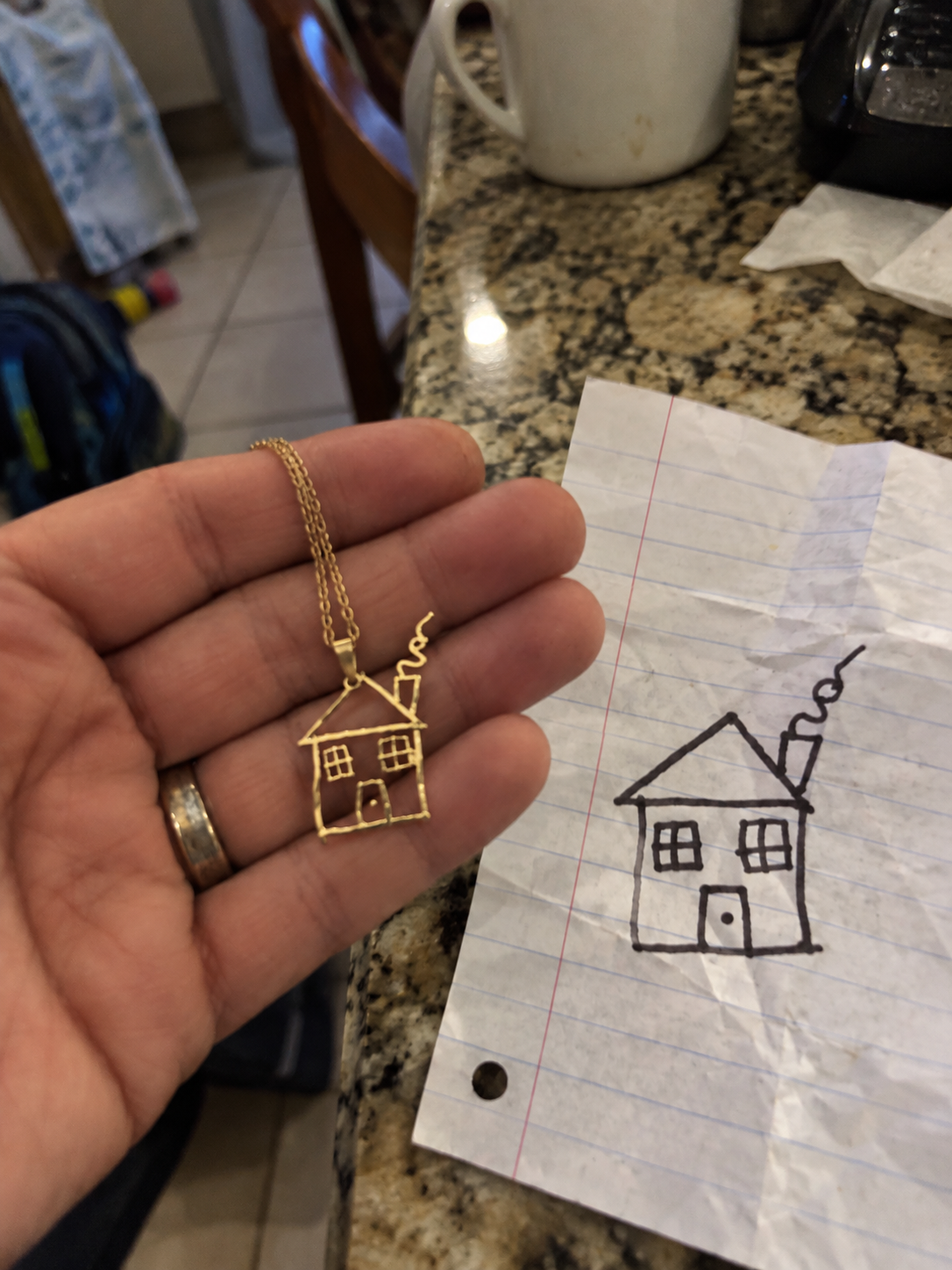Gold house pendant held beside the child's house drawing on notebook paper